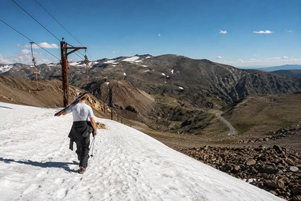 Skier in summer clothing carrying skis to a surface lift on a high mountain snowfield surrounded by snowless terrain