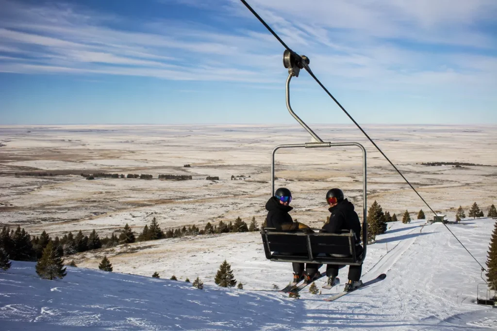 Two skiers riding the Bear Paw double chairlift up with the wide open Hi-Line plains stretching to the horizon in the background