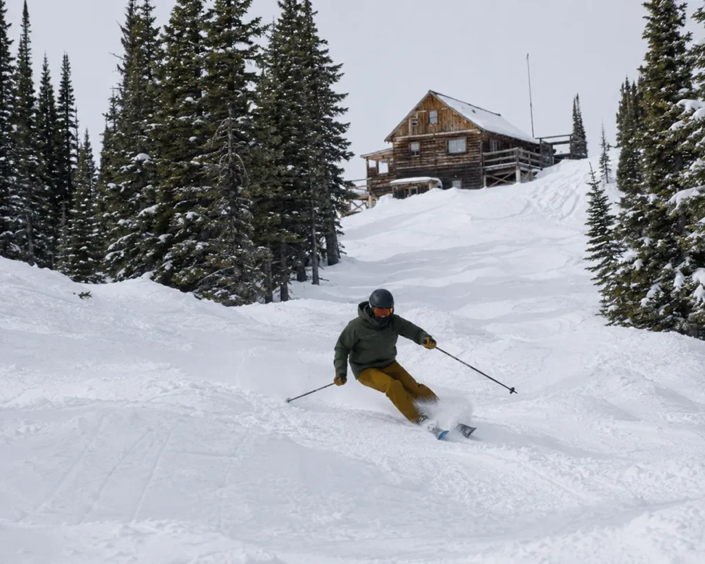 A skier carving down an ungroomed natural snow run at Bear Paw Ski Bowl with a small wooden lodge visible in the distance