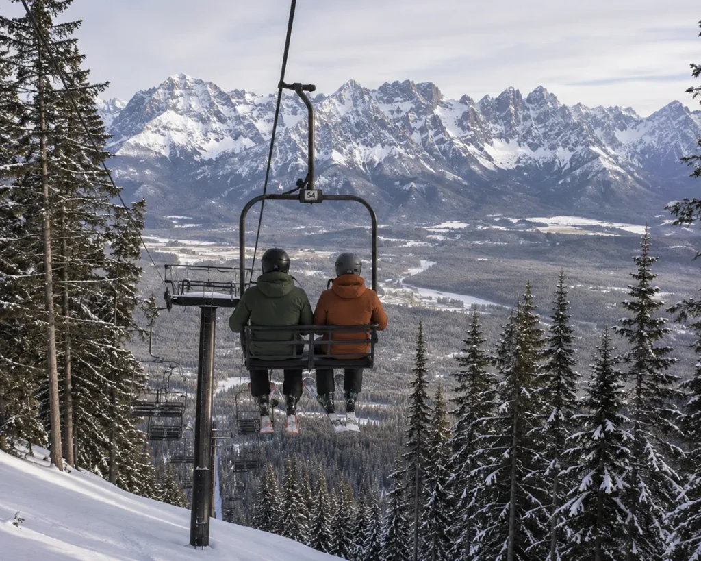 Two skiers riding the Turner Mountain double chair up with snow-covered Cabinet Mountains visible across the valley in the distance