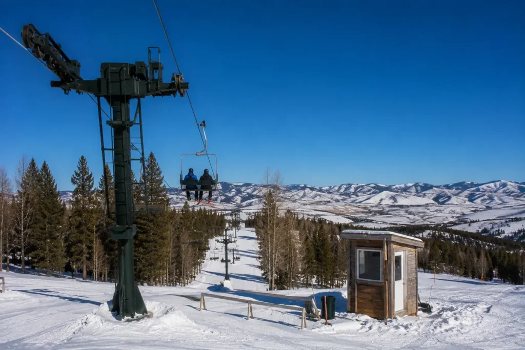 A double chairlift at Bear Paw Ski Bowl with a single skier riding up and the Bear Paw Mountains rising in the background under a blue Montana winter sky