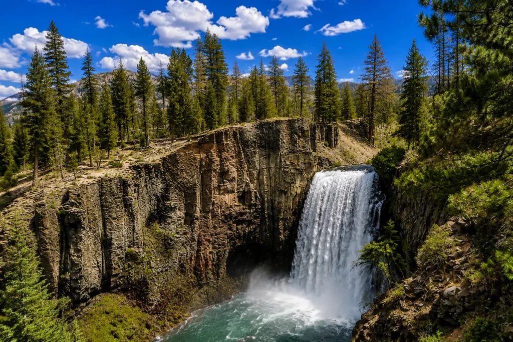 Rainbow Falls in peak spring flow — the Missouri River in full force over the sandstone ledge, spray rising high" Caption: "Rainbow Falls at peak spring flow — Rainbow Dam's spillways overflow and the falls run near their historic volume