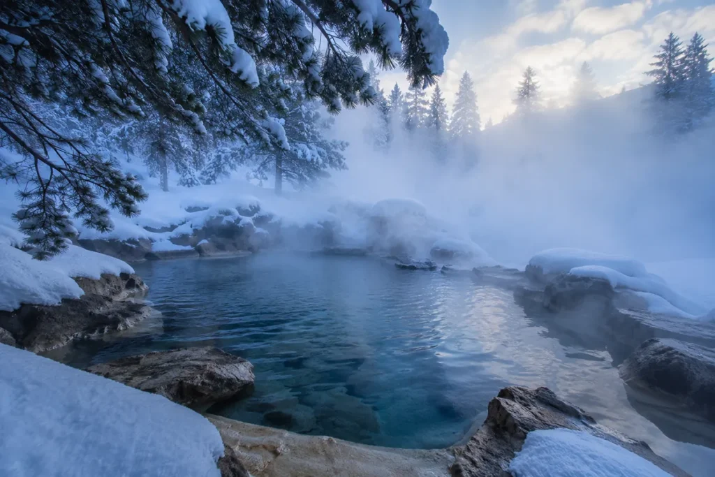 Interior of a steaming Montana hot spring pool surrounded by snow with steam rising into pine trees overhead