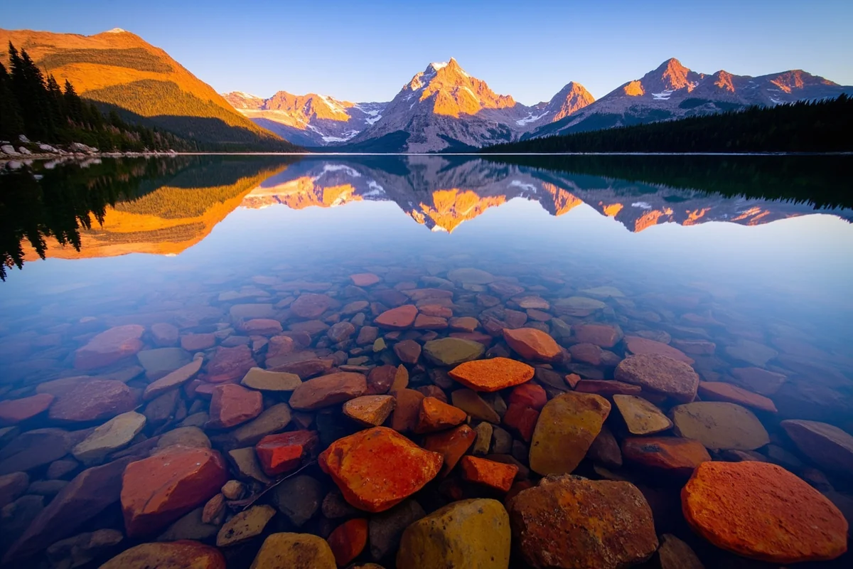 Colorful rocks beneath clear water at Lake McDonald during golden hour with mountain reflections