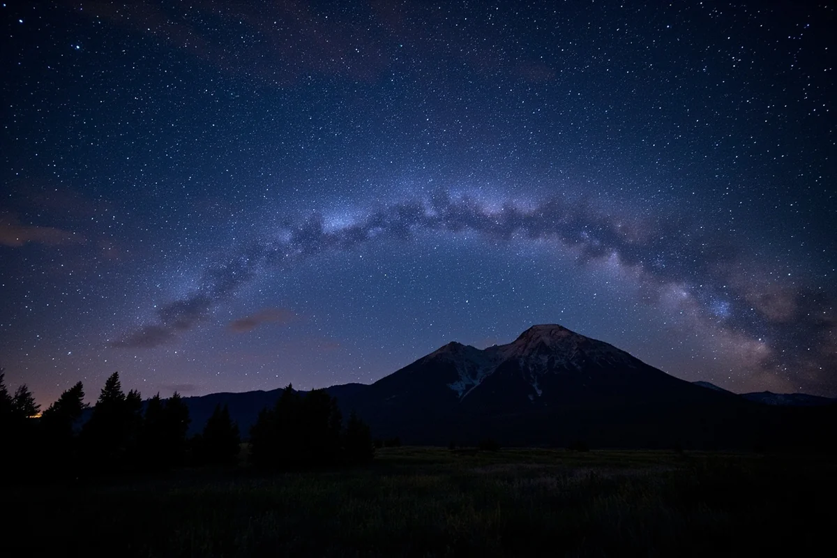 Milky Way arching over a Montana mountain silhouette with foreground meadow
