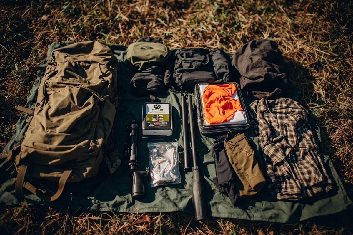 Organized hunting gear laid out on tarp including pack, optics, clothing layers, and camp supplies