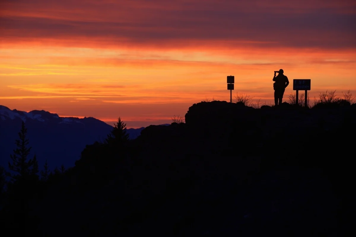 Sunset over Montana mountain landscape with silhouette of hunter glassing from ridgetop, FWP wildlife area sign visible in foreground