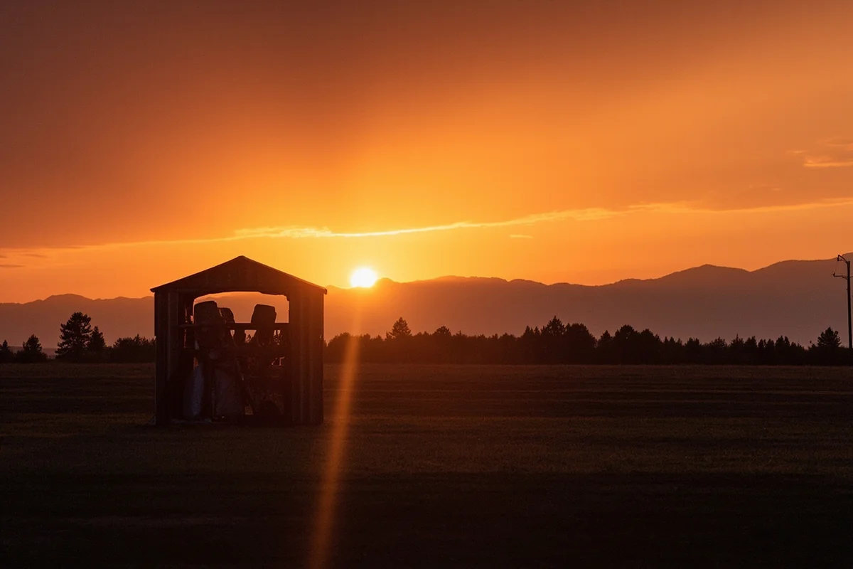 Sunset view over a Montana paintball field with equipment shed in foreground and mountain silhouettes against orange sky