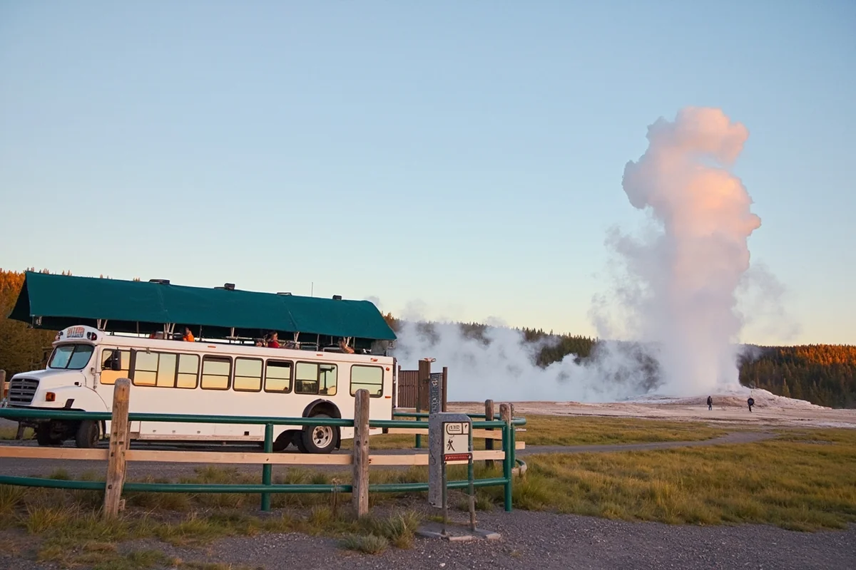 Tour bus parked at Old Faithful viewing area with geyser erupting in background