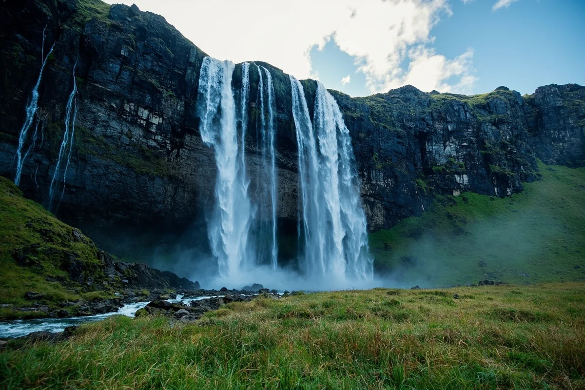 Wide panoramic shot of Silken Skein Falls cascading in multiple ribbons down a dark cliff face with alpine meadows in the foreground
