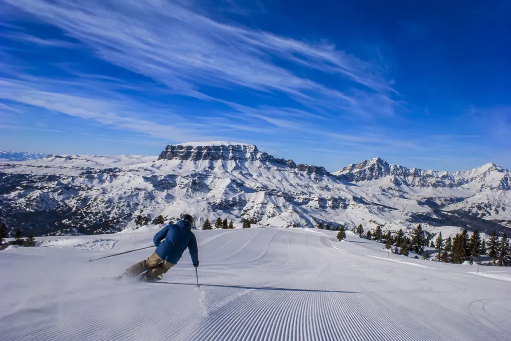 A skier carving a wide turn down the Broadway run at Red Lodge Mountain with the dramatic snow-covered Beartooth Plateau visible in the distance