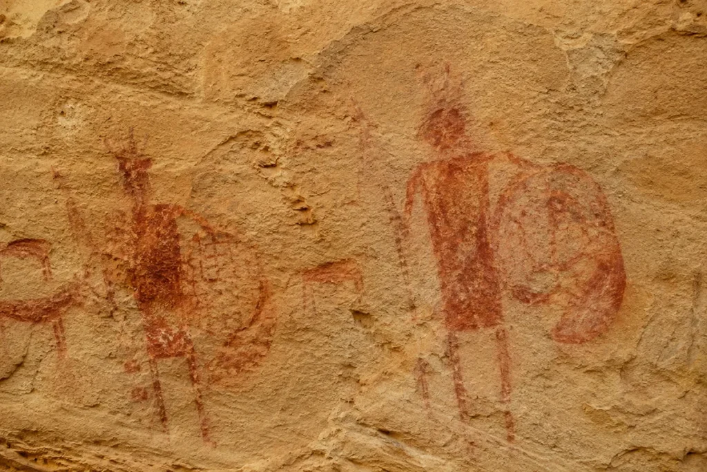 Close-up of red-ochre pictographs on a sandstone wall at Pictograph Cave State Park in Montana, showing faded warrior and animal figures from prehistoric Indigenous artists