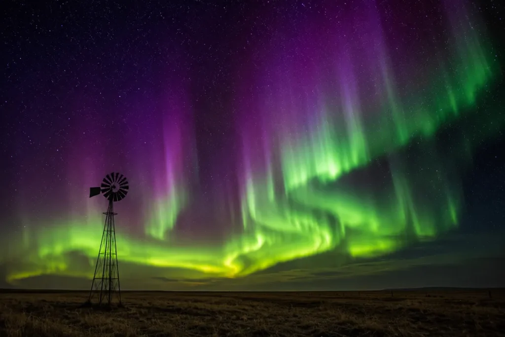 Green aurora borealis dancing across a dark Montana sky above an empty prairie with a single windmill silhouette