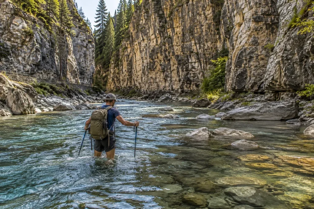 A hiker fording Belt Creek with water reaching mid-thigh inside Sluice Boxes State Park, Montana, with limestone cliff walls rising on either side