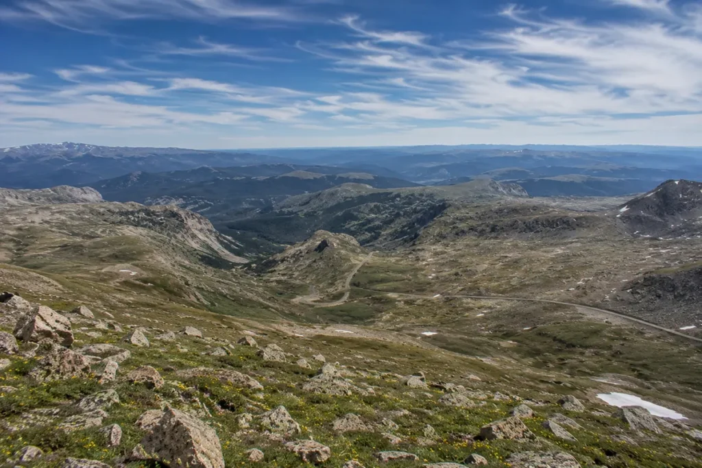 Wide view from the top of Beartooth Pass looking down across the Beartooth Plateau with the highway winding through the valley far below and multiple mountain ranges visible to the horizon