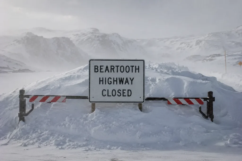 Snow-covered Beartooth Highway sign warning that the road is closed for the winter season with snow drifts visible behind it