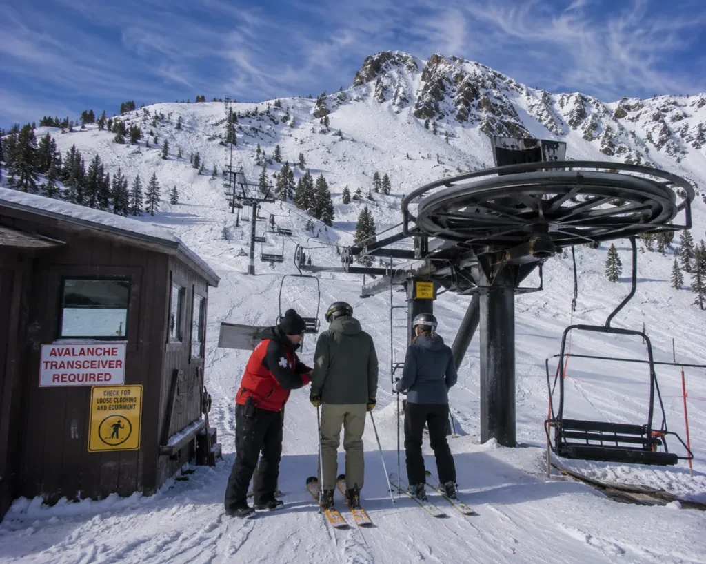 Skiers loading the Schlasman's double chair at Bridger Bowl with the avalanche transceiver check sign visible at the load station and the Ridge rising steeply behind