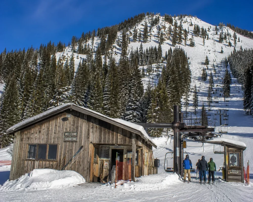 The base lodge and chairlift at Montana Snowbowl with skiers loading the Grizzly Chair under a clear winter sky and the steep TV Mountain face visible behind
