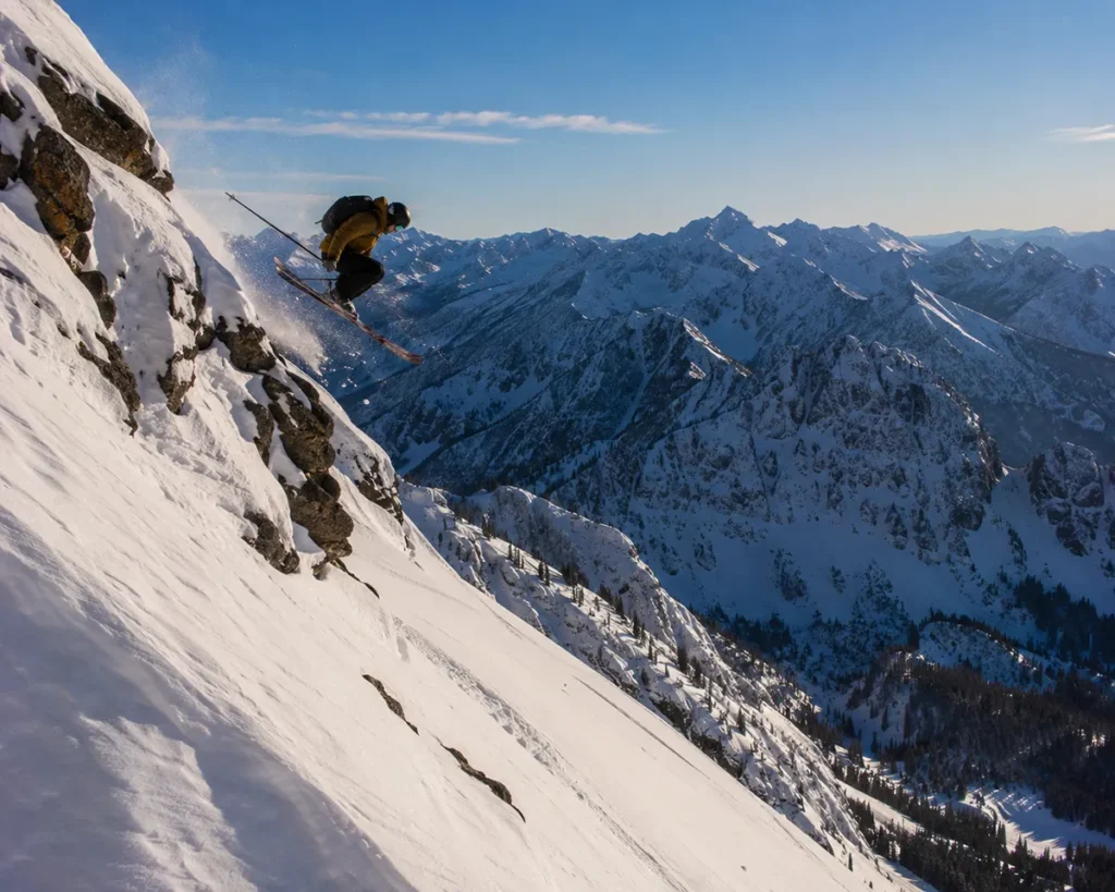 A skier dropping off a small cliff band on the Ridge at Bridger Bowl with steep technical terrain visible below and snowy peaks of the Bridger Range stretching to the horizon