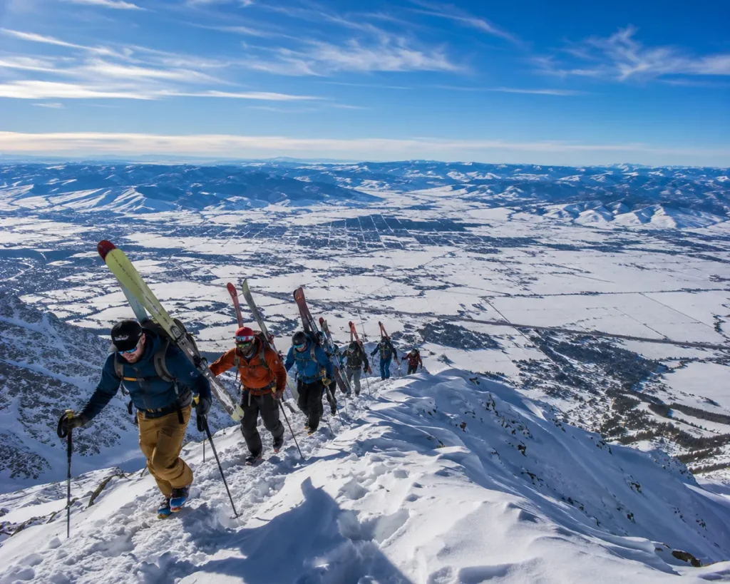 Skiers hiking up the Ridge at Bridger Bowl with avalanche transceivers visible and the Gallatin Valley spread out far below under a deep blue Montana winter sky