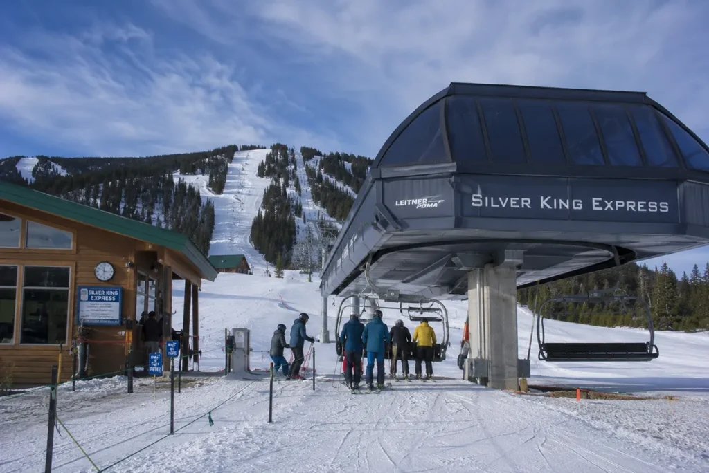 The base lodge area at Whitefish Mountain Resort with the Snow Ghost Express six-pack chairlift loading skiers under a clear sky and the historic Hellfire run visible behind