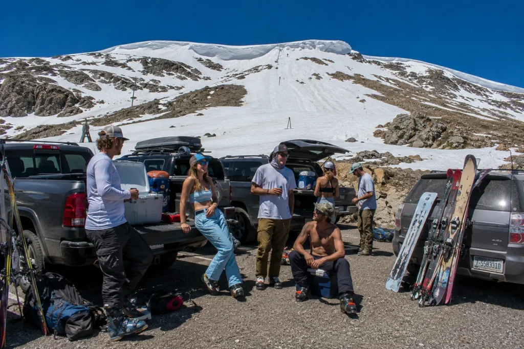 Skiers tailgating in the Beartooth Basin parking lot with skis and gear visible against pickup trucks and the high alpine ski terrain rising behind
