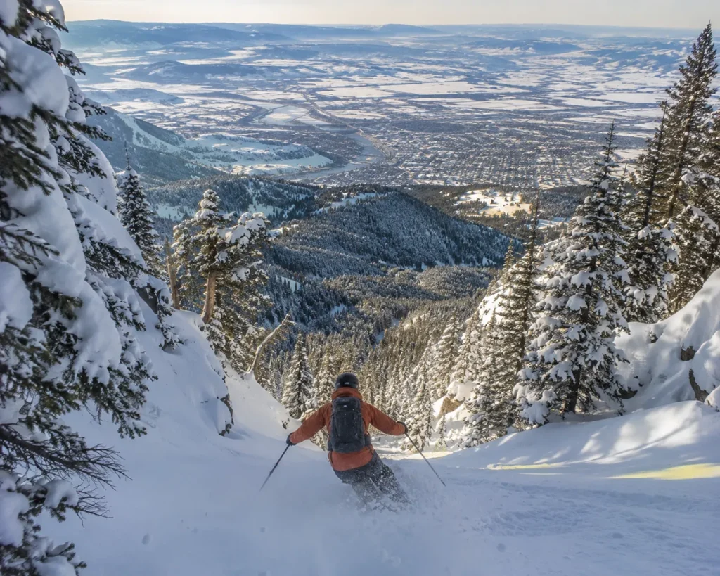 A skier dropping into the steep TV Mountain terrain at Montana Snowbowl with the Missoula valley visible far below in the distance
