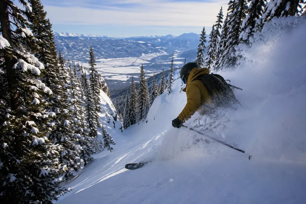 A skier dropping into a steep tree-lined chute off the Challenger lift at Big Sky Resort with deep powder visible underfoot