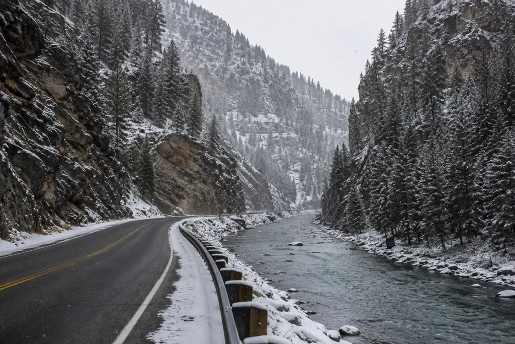 The Gallatin River canyon road in winter with US-191 running alongside the snow-covered river and sheer rock walls rising on both sides
