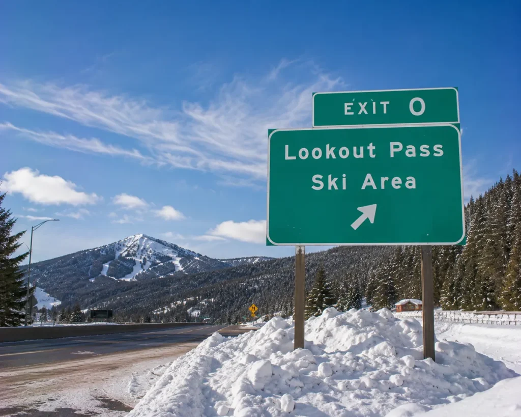An Interstate 90 highway sign for Exit 0 with snow-capped mountains visible behind and the Lookout Pass ski area facilities partially visible
