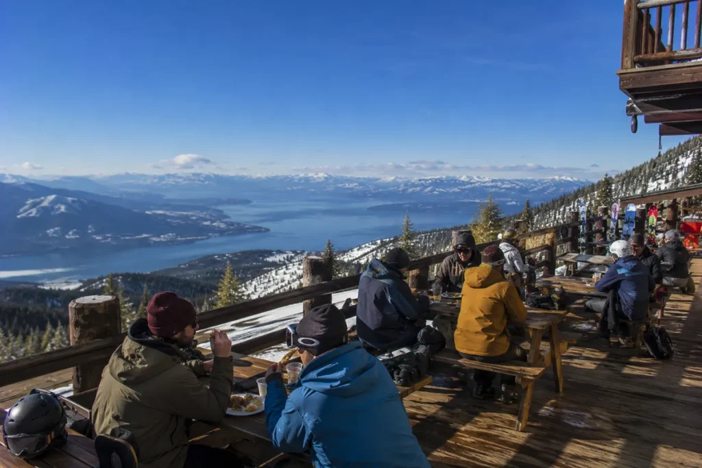 Mid-day view from the deck of the Blacktail Mountain summit lodge with skiers visible enjoying lunch and the vast snow-covered Flathead Valley spread out below