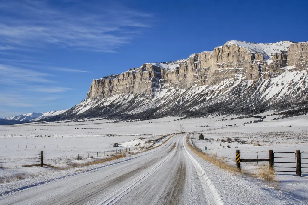 Snow-covered Teton Canyon Road winding west from Choteau toward the dramatic Rocky Mountain Front cliffs visible in the distance