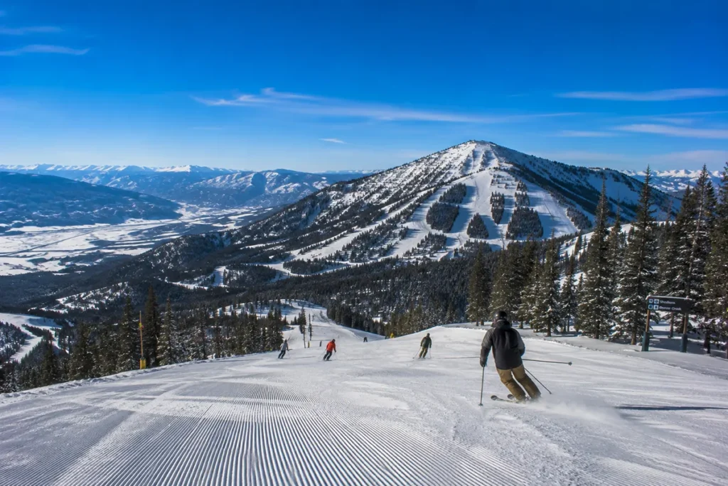 View from the top of Andesite Mountain at Big Sky Resort looking across to Lone Peak with skiers carving down a wide groomed run in the foreground