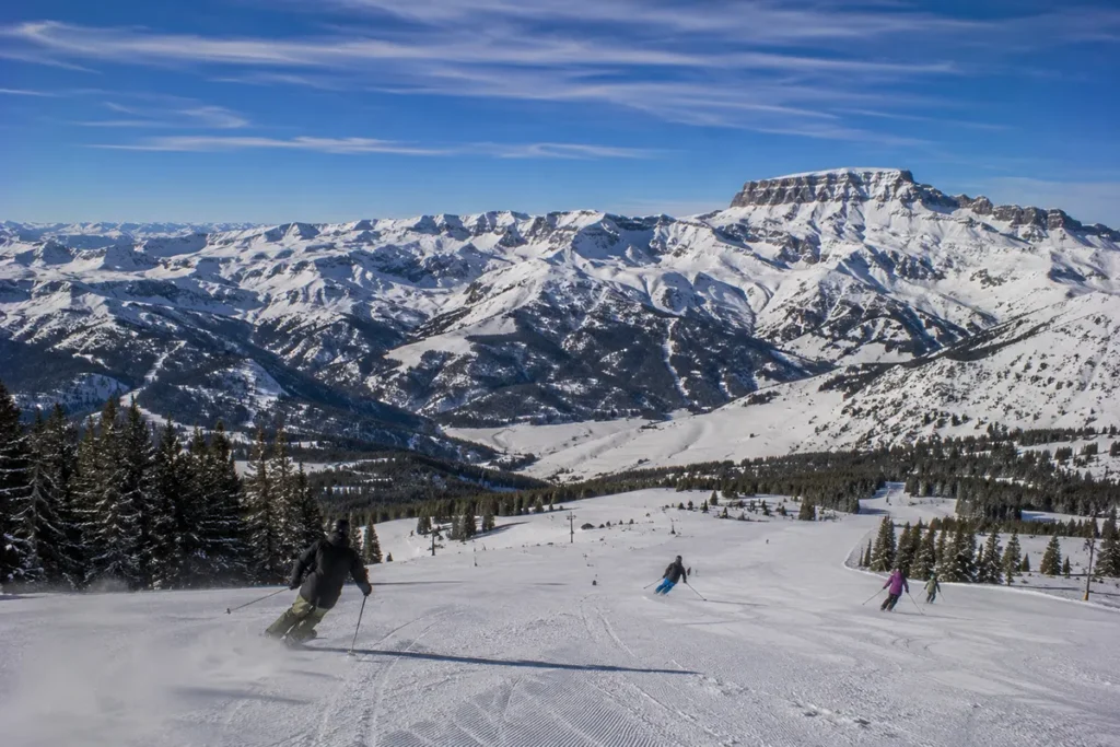 View from the top of Red Lodge Mountain Resort looking out across the snow-covered Beartooth Range with skiers visible carving down a wide groomed run in the foreground