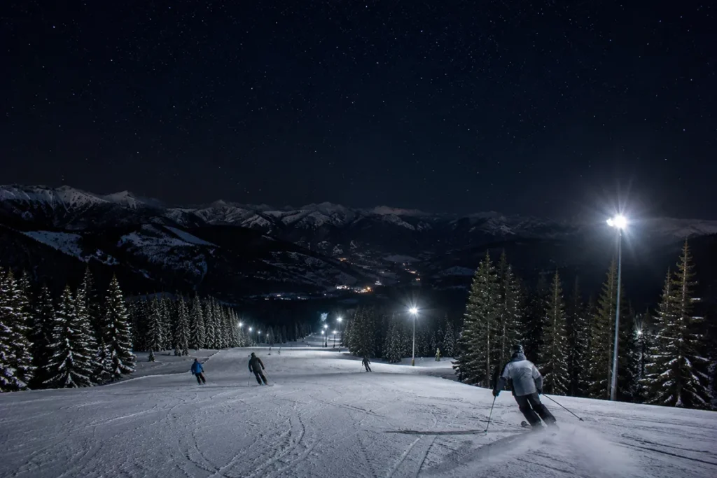 Night skiing at Great Divide Ski Area with skiers descending an illuminated run under floodlights and a starry sky visible above