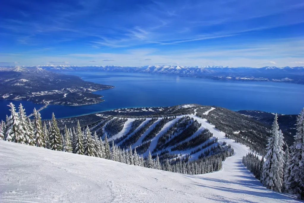 Panoramic view from the top of Blacktail Mountain looking down across snow-covered slopes to the deep blue waters of Flathead Lake with the Mission Mountains rising in the distance on a clear winter day