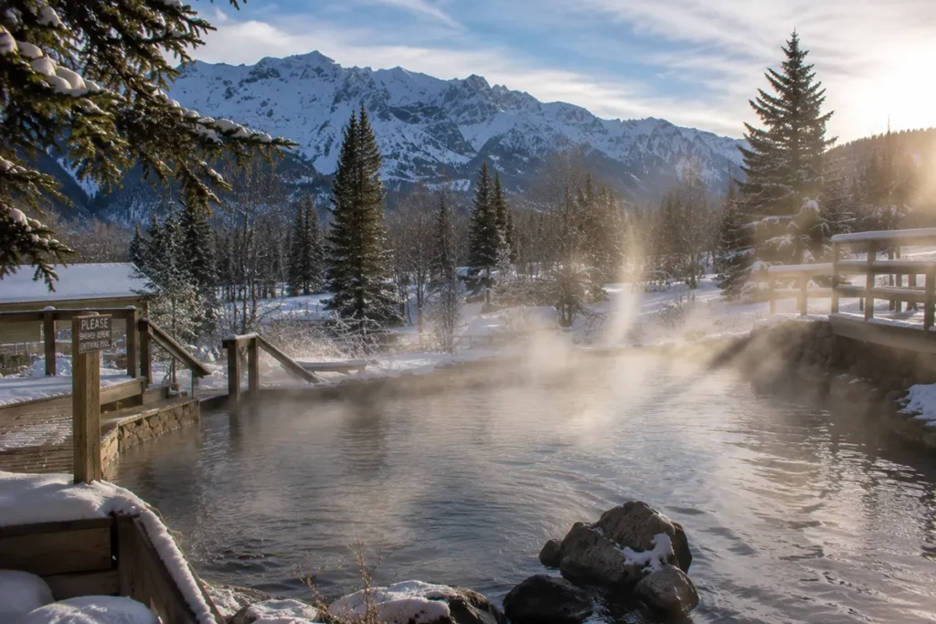 View of Elkhorn Hot Springs steaming pool with snow on the surrounding deck and mountains visible behind on a sunny winter day