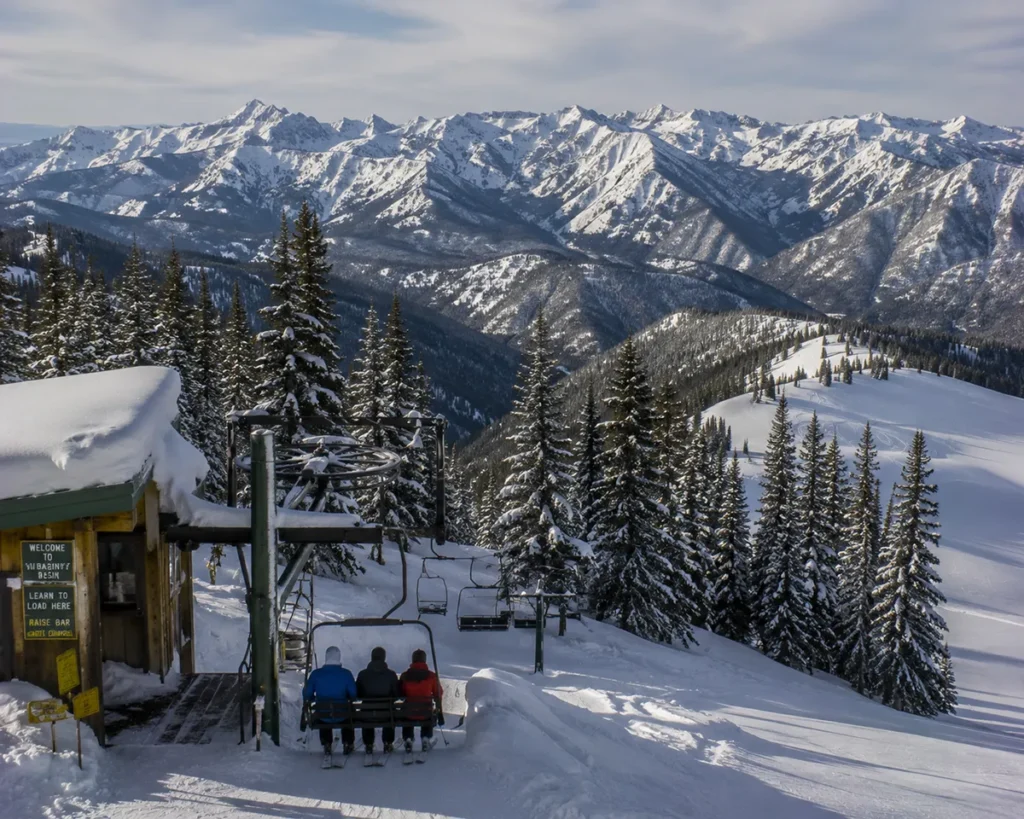 Skiers riding a vintage double chairlift at Lost Trail Powder Mountain with deep untracked powder visible on the slope beside them and the Continental Divide ridge stretching across the background