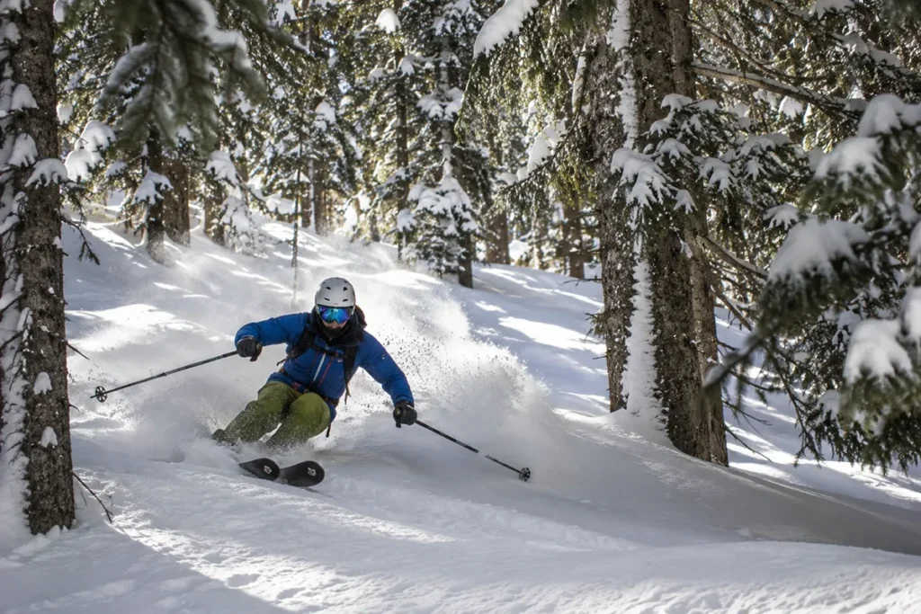 A skier weaving between widely spaced evergreens in a designated glade zone at Montana Snowbowl with fresh powder spraying from skis
