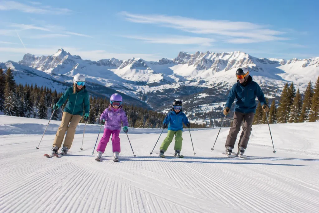 A family of four skiing together down a wide groomed run at Red Lodge Mountain with the parents on either side of two children on a sunny winter morning