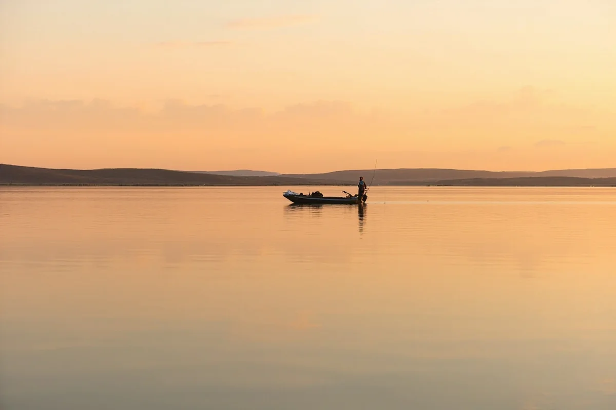 Angler in a small fishing boat on Fort Peck Reservoir at sunrise, with pale orange sky reflecting on calm water and badland banks in the background